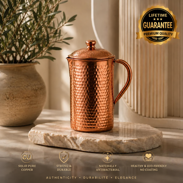 Hammered copper water carafe with lid and handle on a marble slab, styled with a potted plant