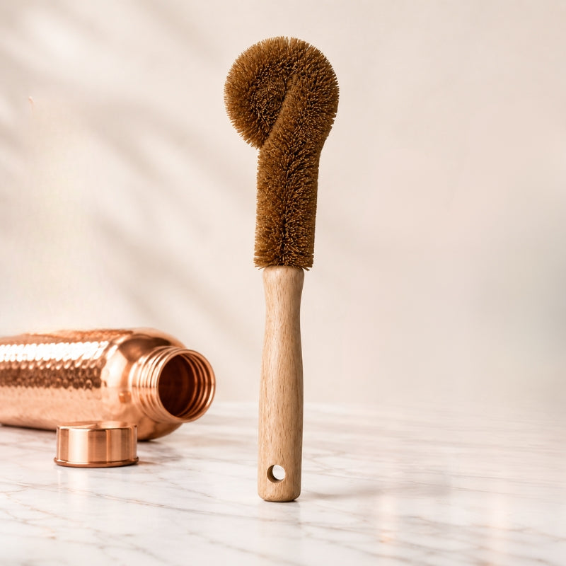 Natural coconut-fibre cleaning brush with wooden handle standing next to a copper water bottle