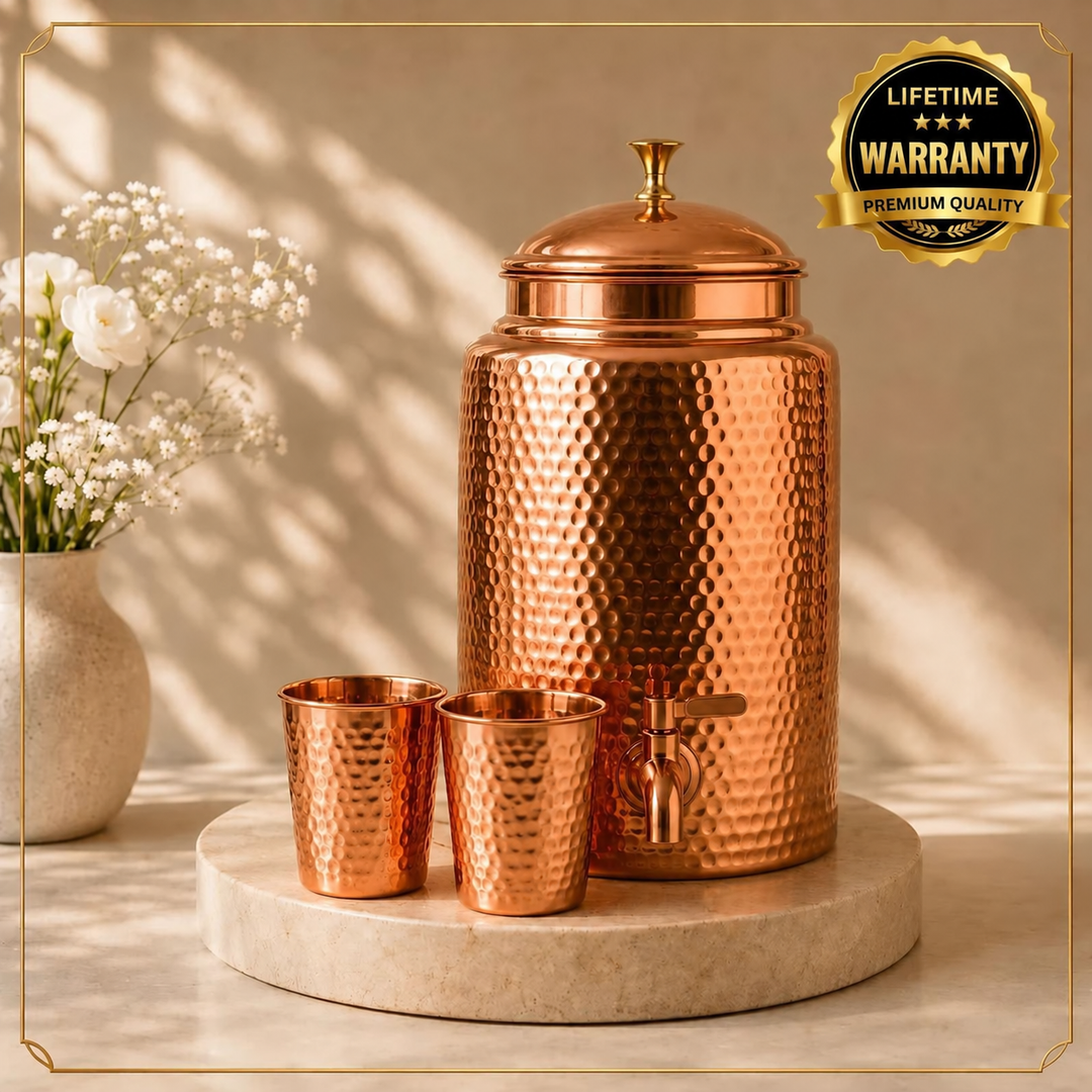 Hammered copper water dispenser with tap and lid, two matching copper cups, on a marble stand with flowers
